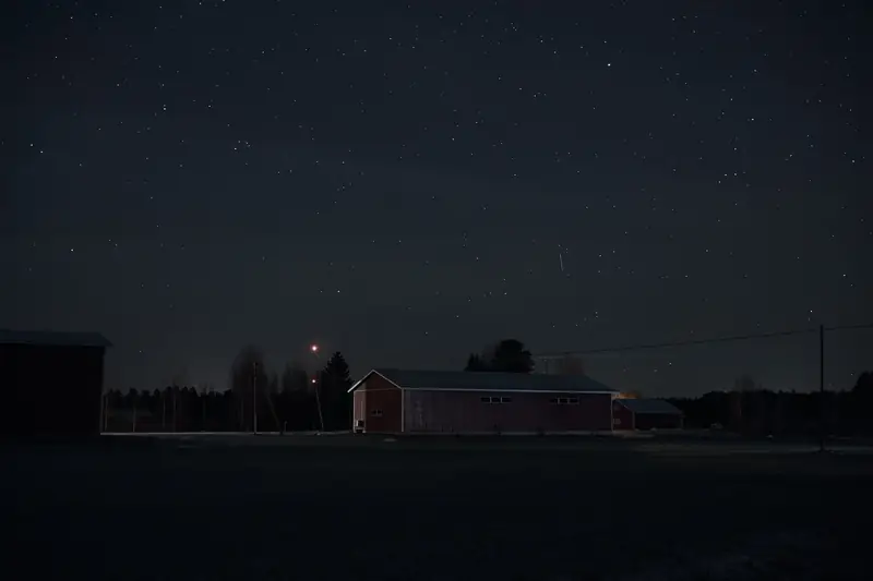 A dark landscape photograph of a large, rectangular farm building silhouetted against a vast, starry night sky. The scene is deeply shadowed, with only a few faint lights visible in the distant background.